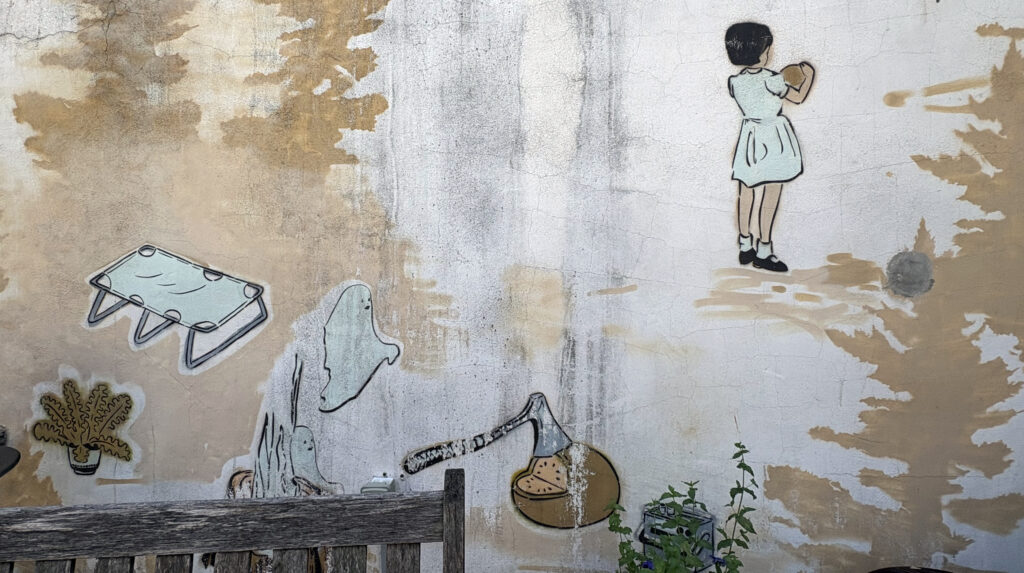 Mural by Mando Marie with a girl, ghosts, a cot, a potted plant and an axe chopping a melon.